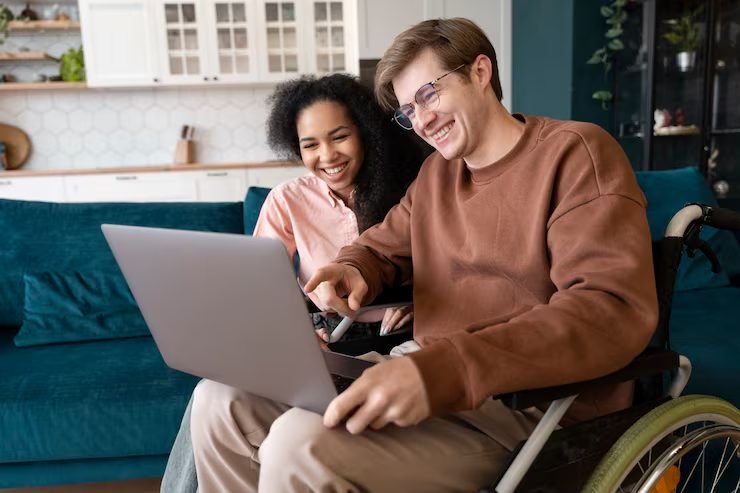 A young man using a wheelchair, relaxed at home with a friend, looking at a laptop together